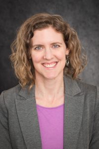 A photo of RPI Partner Clare Levin. Clare has curly hair, is wearing a grey suit jacket with a purple top and is similing.